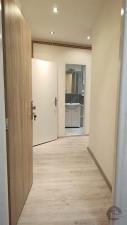 Úzká chodba s dřevěnou podlahou, moderní osvětlení, bílé stěny, dveře vedou do koupelny s dlažbou a umyvadlem, dobrý stav.