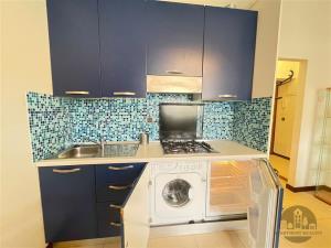 Moderní kuchyň s modrými skříněmi, mozaikovým obkladem a vestavěnými spotřebiči, jako je pračka a plynový sporák, na světlé dlažbě.